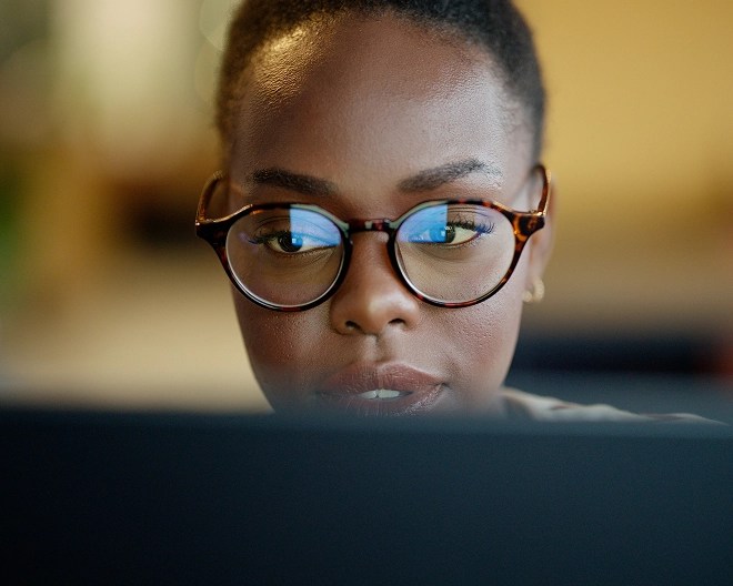 Close-up of a person slightly smiling as she works at a computer, her work reflected in her glasses.