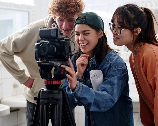 Three young media professionals stand as they review photos on a camera sitting atop a tripod.