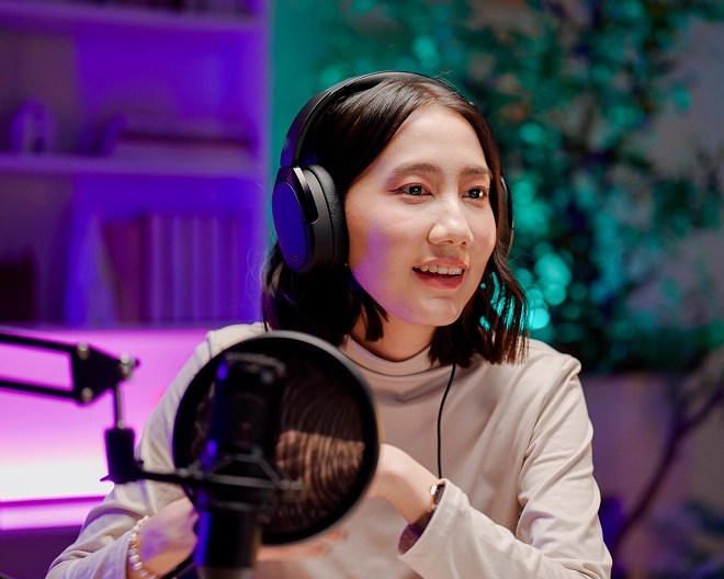Podcaster wearing headphones looks out of frame to her guest in a room lit in pinks, purples, and teals.