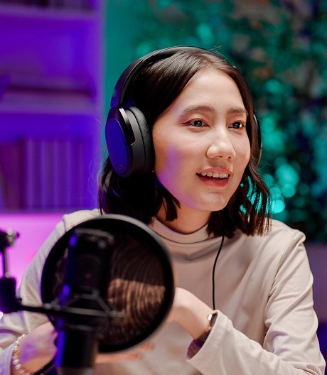 Podcaster wearing headphones looks out of frame to her guest in a room lit in pinks, purples, and teals.