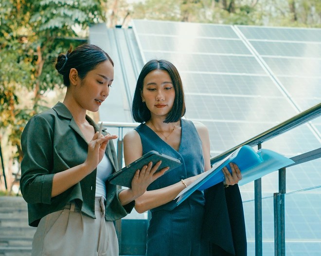Two sustainability experts stand in front of a solar panel, looking at a tablet and an open folder.