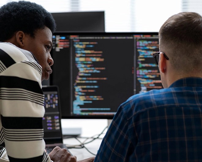 A pair of male colleagues collaborate at a desk, reviewing lines of code on dual computer monitors in a bright office setting.