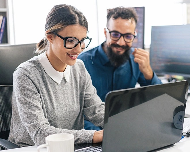 A pair of colleagues collaborate on an open laptop in a bright office.