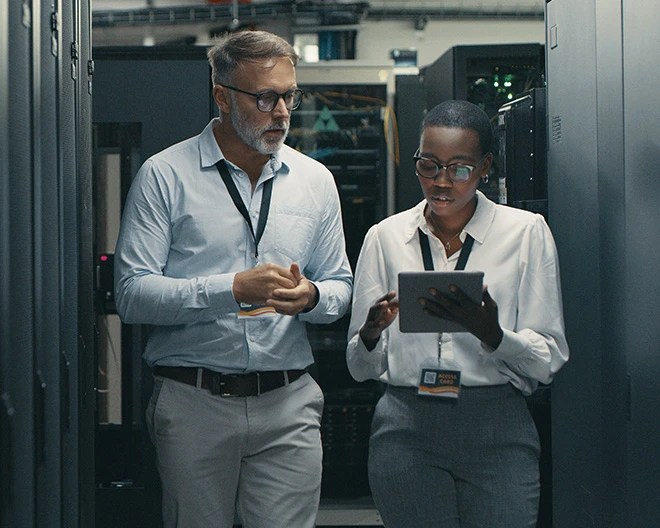 Two IT professionals walk through a tech room, one holding a tablet.