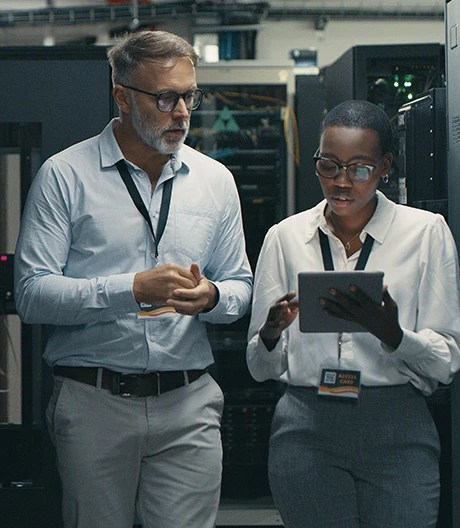 Two IT professionals walk through a tech room, one holding a tablet.