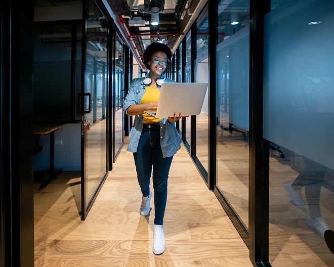 Smiling tech professional wearing glasses, a yellow top, denim jacket, and headphones around her neck, walking through an office corridor while holding an open laptop.