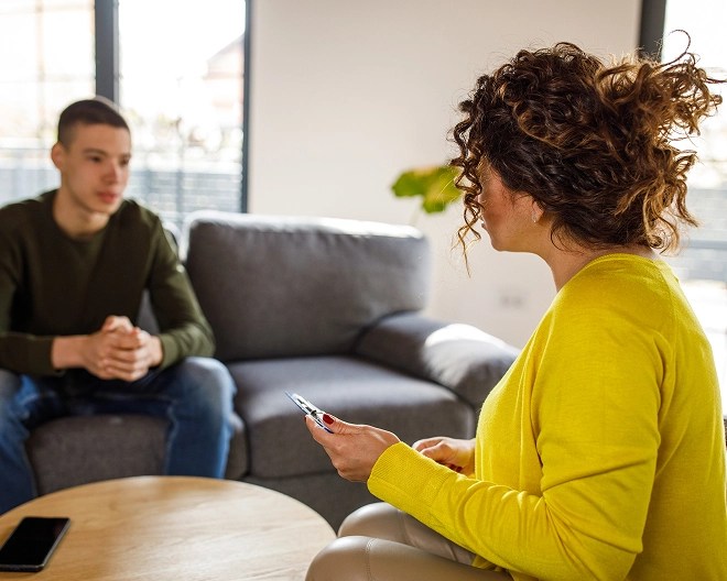 Therapist in a yellow sweater attentively listening to a young client seated on a couch during a therapy session.