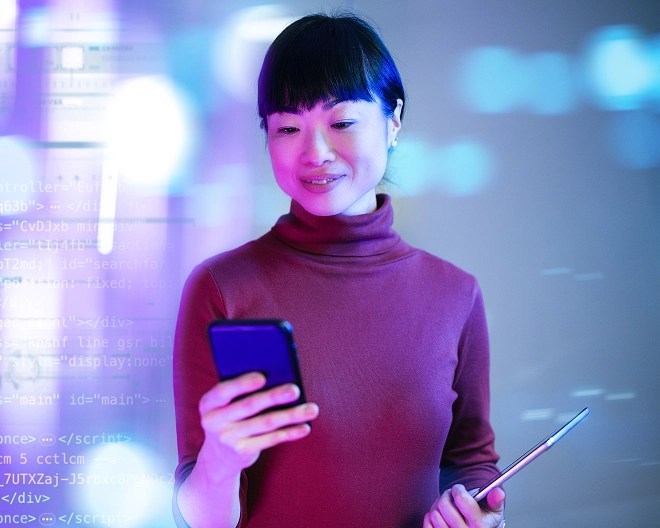 Tech professional wearing a turtleneck, smiling while looking at her phone, with a presentation projected behind her in vibrant blue, purple, and pink hues.