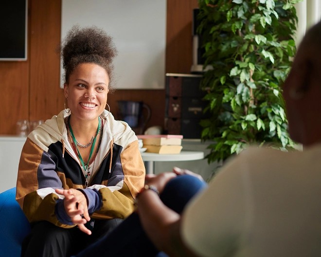 Patient with her hair up and wearing a sports jacket smiles during a therapy session, while the therapist listens attentively with legs crossed.