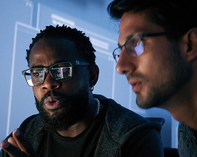 Close-up of two data scientists analysing information on a computer out of frame, data projected on the wall behind them.