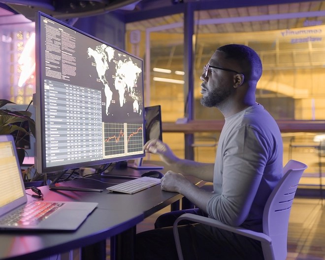 Data scientist analyzing numbers, charts, and a global map on a desktop computer at night, with the screen casting a soft glow in a dark room.