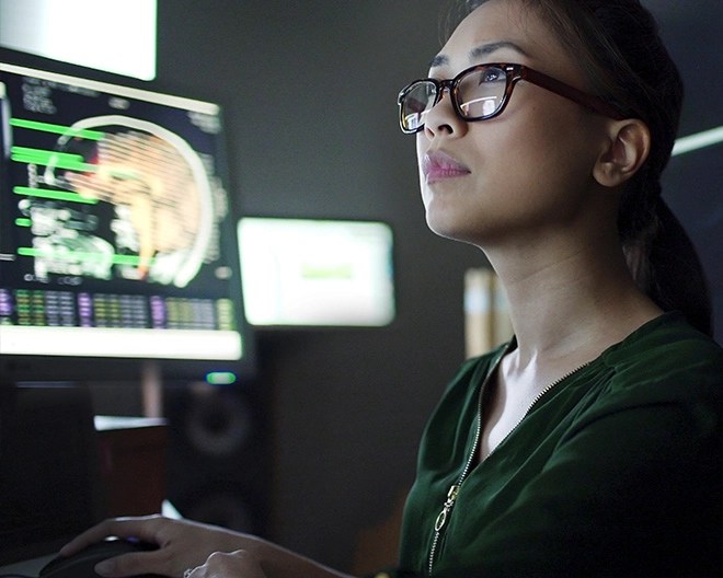 A woman wearing glasses reviews a brain scan on a computer monitor in a darkened room, focused on the screen.
