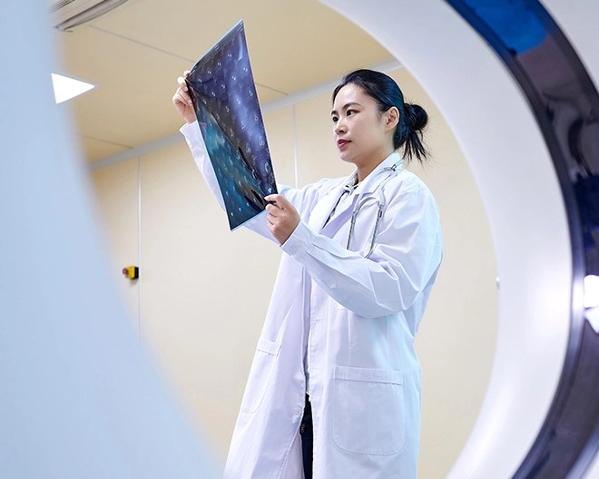 A female doctor in a white lab coat examines a brain scan held up to the light, standing beside medical imaging equipment.