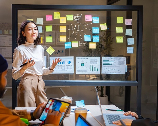 Woman presenting AI concepts on a transparent board covered with sticky notes and data charts, in a modern office meeting room.