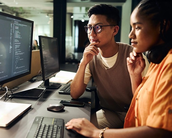 Two colleagues review code on a desktop computer in a bright office.