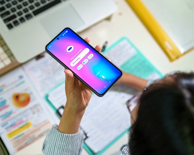 Close-up of a person holding a smartphone displaying a voice assistant AI app interface, with study materials and a laptop in the background.