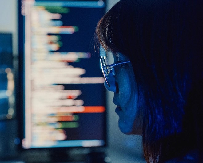 Woman focused on a computer screen displaying lines of code, illuminated by the blue light from the monitor in a dark room.
