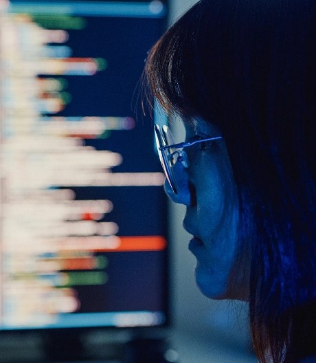 Woman focused on a computer screen displaying lines of code, illuminated by the blue light from the monitor in a dark room.