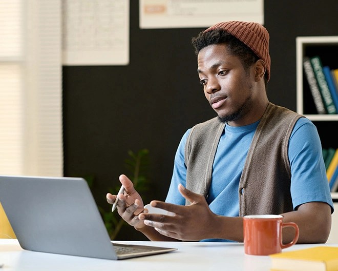 Online student wearing a light blue t-shirt, open vest, and orange knit hat communicates on his laptop in an office.