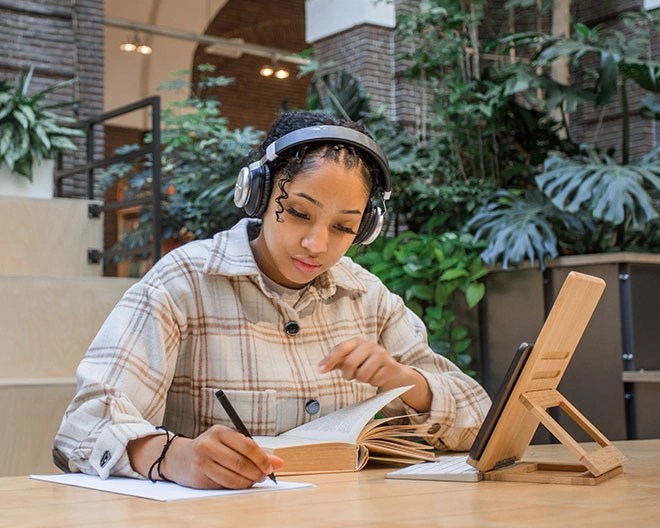 A young woman wearing headphones studies in a modern indoor space with lush greenery, taking notes from an open book while referencing a tablet propped on a wooden stand.