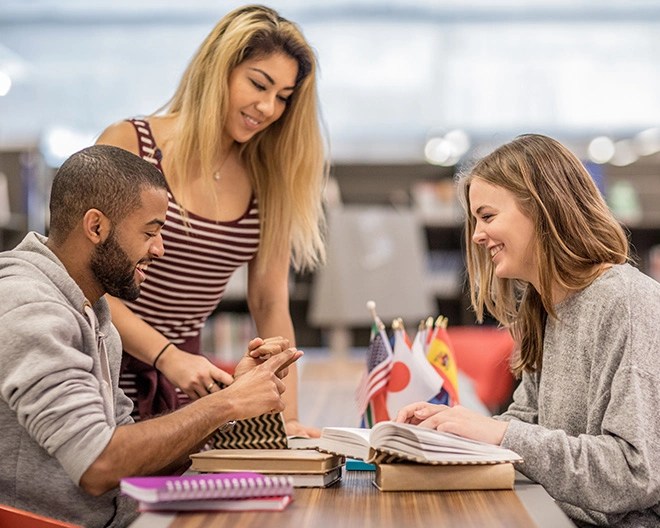Three smiling students meet up at the library to talk linguistics, their table filled with notebooks and flags.