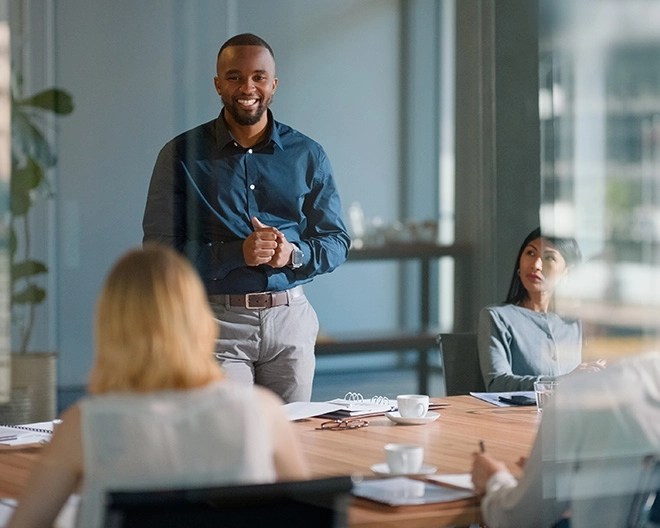 A businessman in business casual attire stands at an office table, presenting to his colleagues during a meeting.
