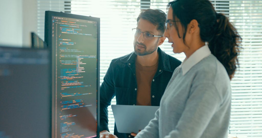 Two individuals collaborating in front of a computer screen displaying lines of code, likely in JavaScript or Python.