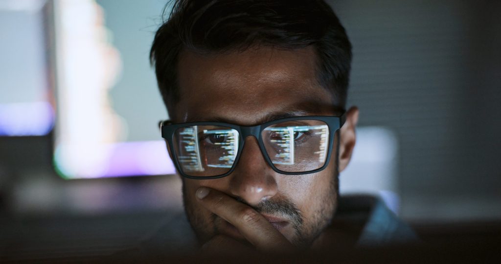 Person wearing glasses, seated in front of a computer screen displaying colourful graphics, with a blurred background.