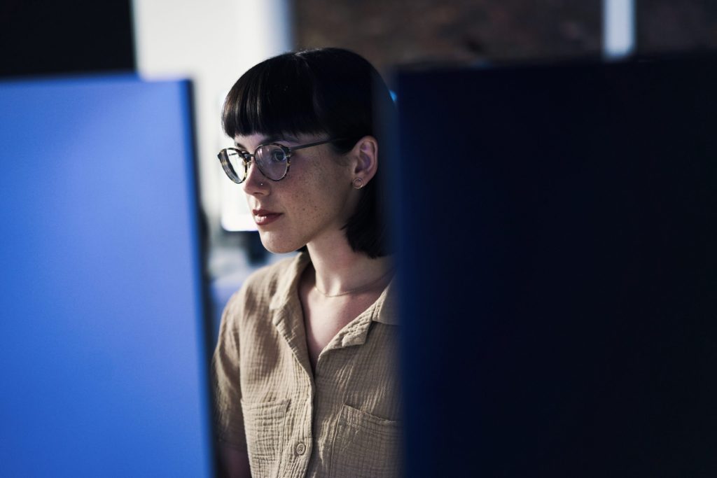 Data scientist with short hair, glasses, and a tan button-up works behind a computer in a dimly lit office.