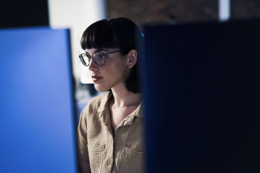 Data scientist with short hair, glasses, and a tan button-up works behind a computer in a dimly lit office.