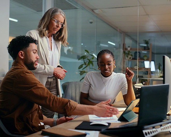 Three colleagues discuss a tech-related topic in a modern office building, their meeting table filled with papers and laptops.