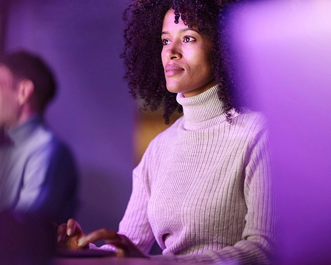 A woman in a white knit turtleneck works at her computer, illuminated by soft purple lighting.
