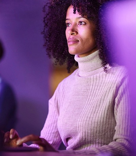 A woman in a white knit turtleneck works at her computer, illuminated by soft purple lighting.