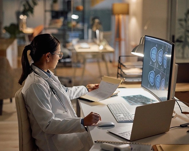 A doctor in a white coat and stethoscope sits at a desk in a medical office, reviewing charts and brain scan images on a monitor.