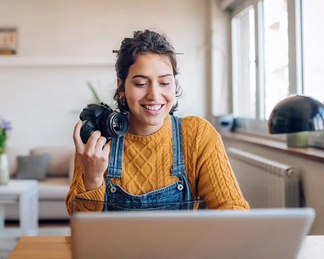 Person wearing a mustard-coloured knit sweater and overalls, holds a camera and smiles while she works on the computer at home.