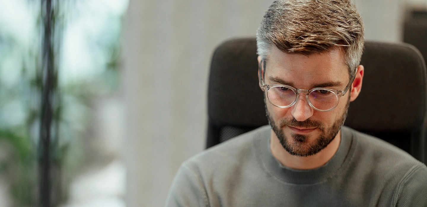 A man wearing glasses and a grey jumper looking at a computer screen.