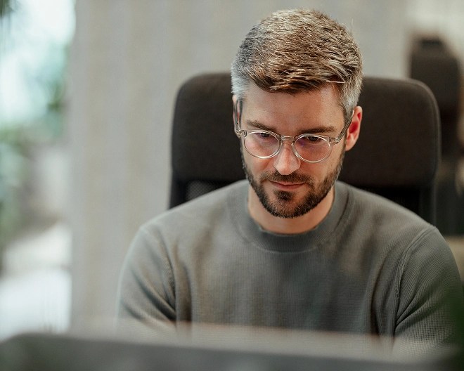 A man wearing glasses and a grey jumper looking at a computer screen.