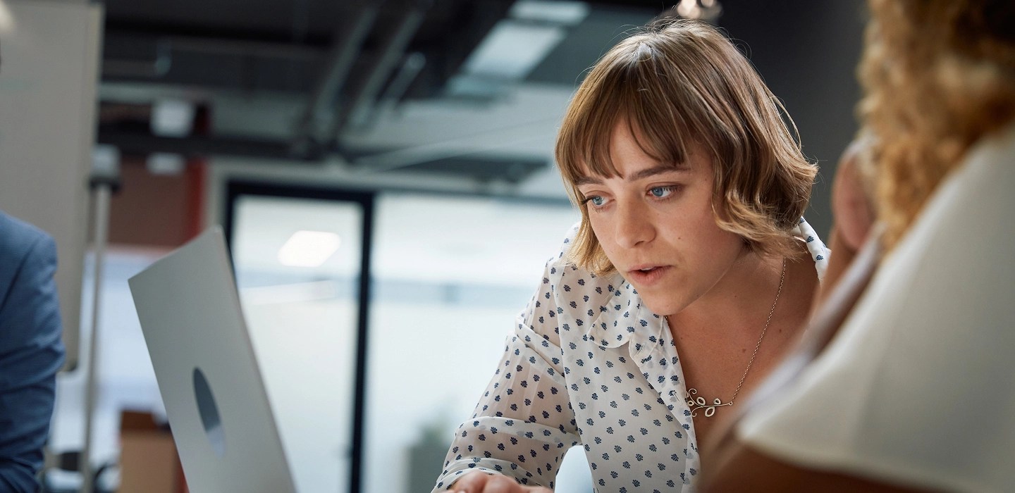 A woman wearing a white shirt with blue patterns and a silver necklace looking at a laptop with colleagues.