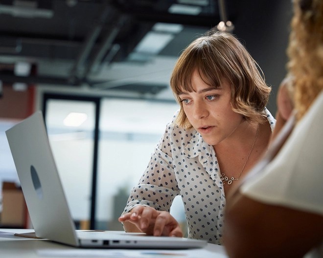 A woman wearing a white shirt with blue patterns and a silver necklace looking at a laptop with colleagues.
