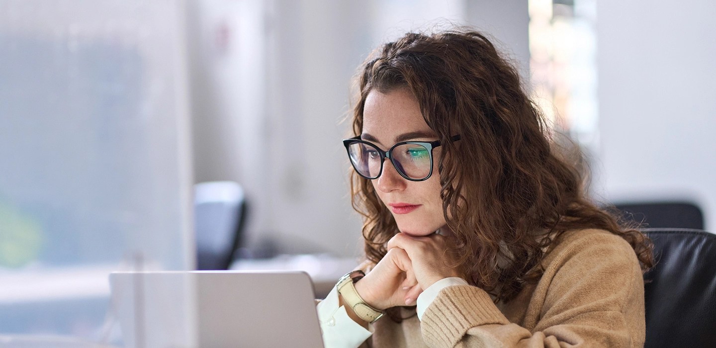 A student wearing glasses and a light brown jumper layered over a white shirt focusing on a laptop screen.