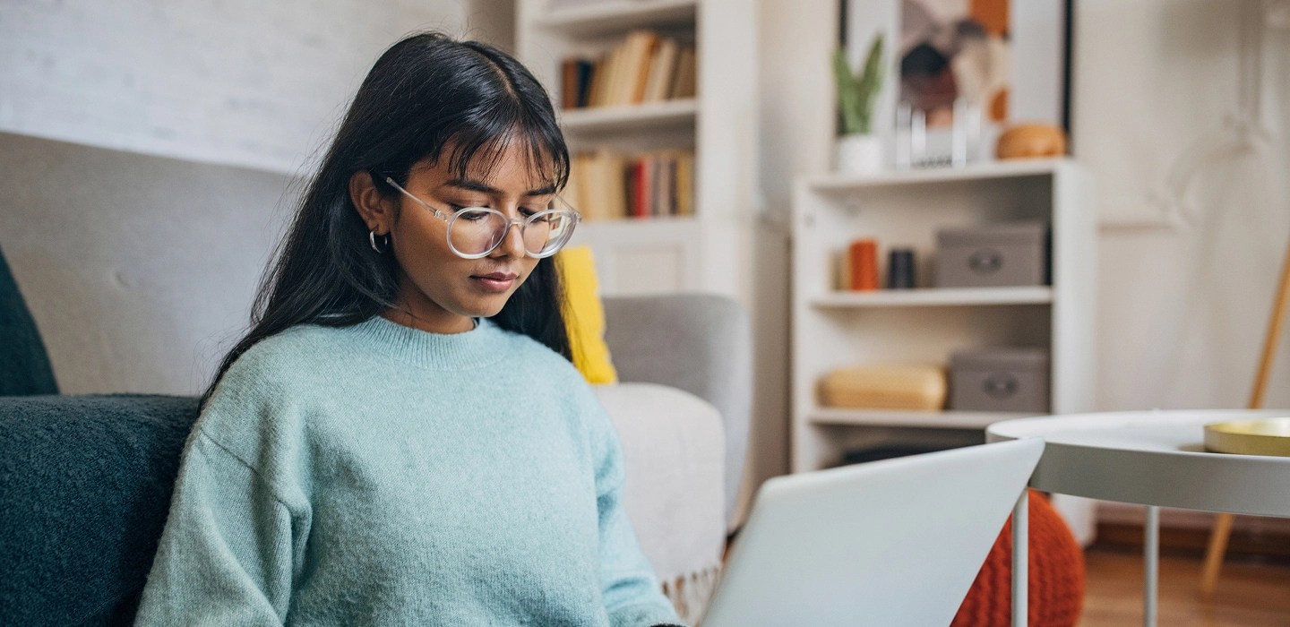 A student sitting on a sofa in a blue sweater, typing on her laptop with a pillow on her lap.