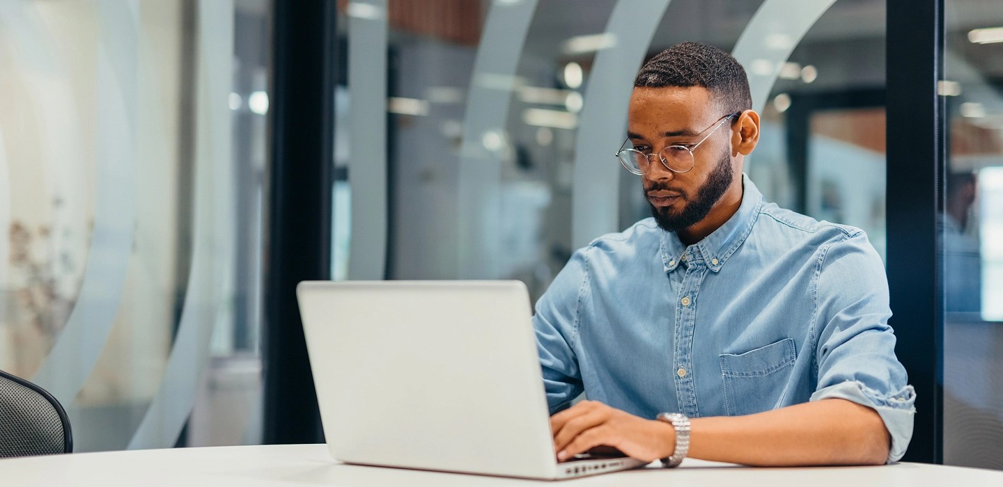 A man in a blue button-up shirt and glasses typing on his laptop in an office.