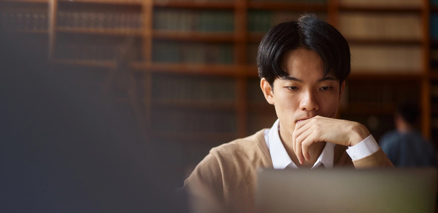 A student wearing a light brown jumper over a white shirt, looking at a laptop screen.