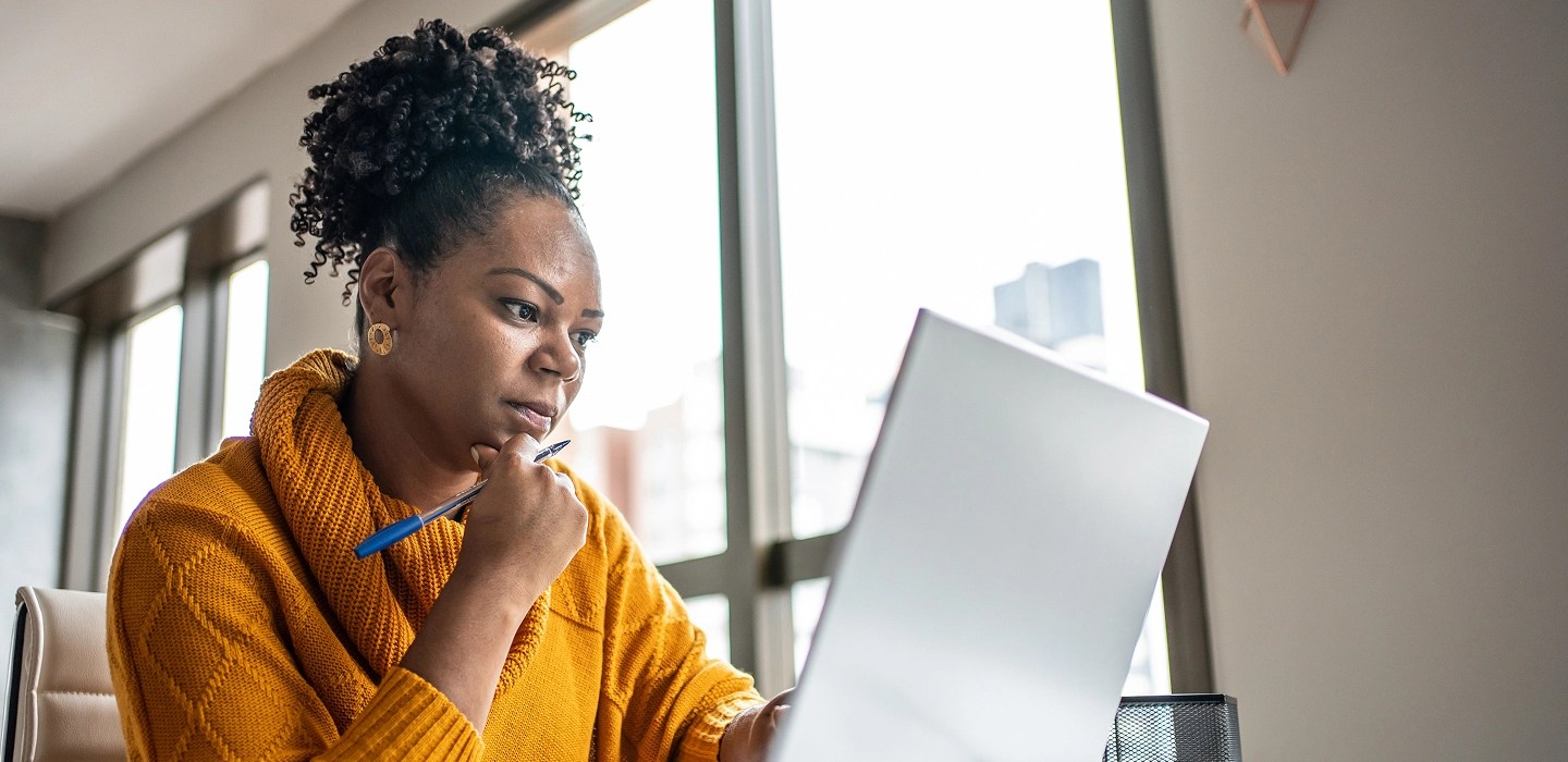 A woman in a yellow sweater holding a pen and looking at her laptop.