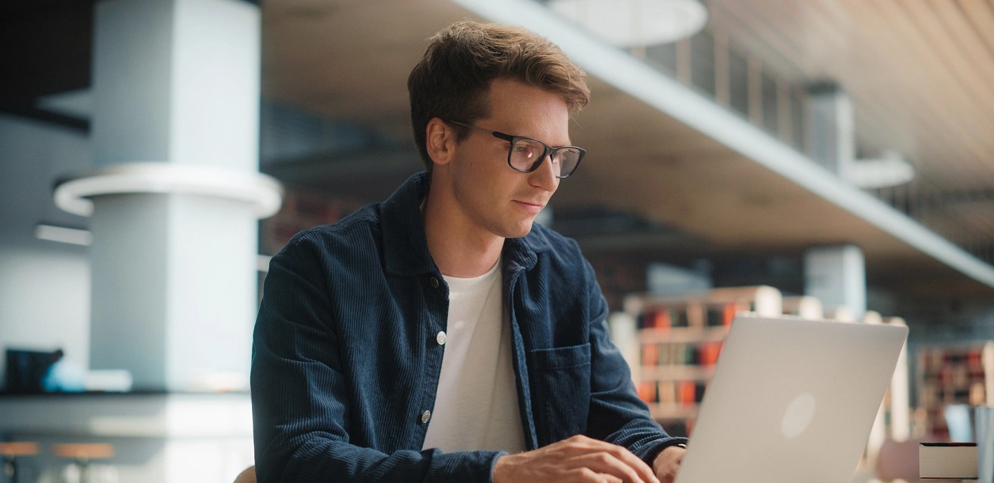 A student wearing glasses typing on his laptop at a library desk, with a notebook nearby.