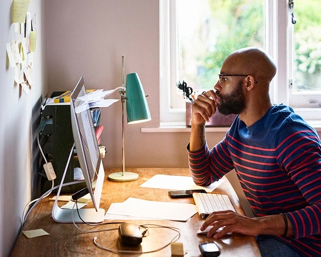 A bald man wearing glasses and a blue and red striped jumper, looking at a computer screen in an office.
