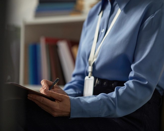 Person in a blue shirt with a white lanyard and ID badge writes on a clipboard, seated in front of shelves filled with books and miscellaneous items.