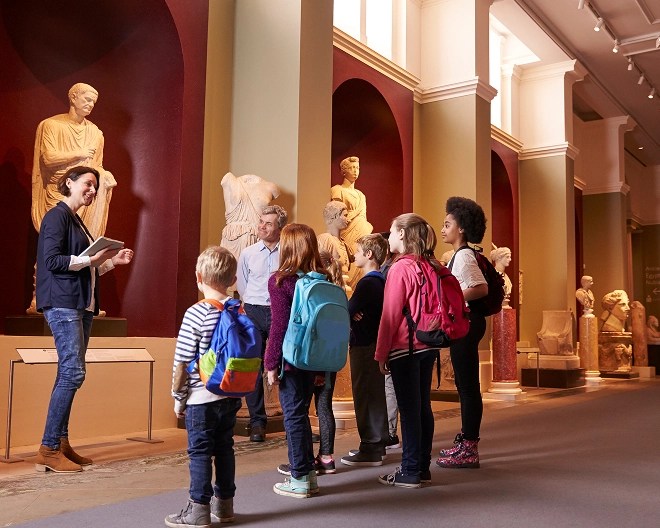 Museum guide holding a tablet leads a group of children and adults on an educational tour of classical sculptures displayed in ornate niches.