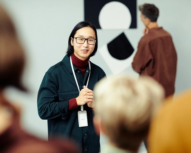 Art museum tour guide wearing a navy jacket, dark red button up, and glasses, speaks to a group of people. A black and white painting is hung behind them.