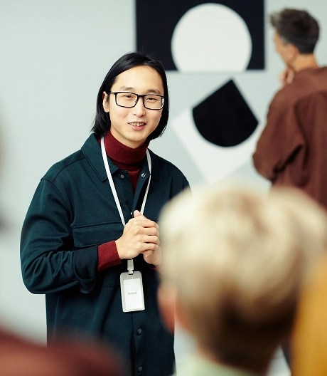 Art museum tour guide wearing a navy jacket, dark red button up, and glasses, speaks to a group of people. A black and white painting is hung behind them.